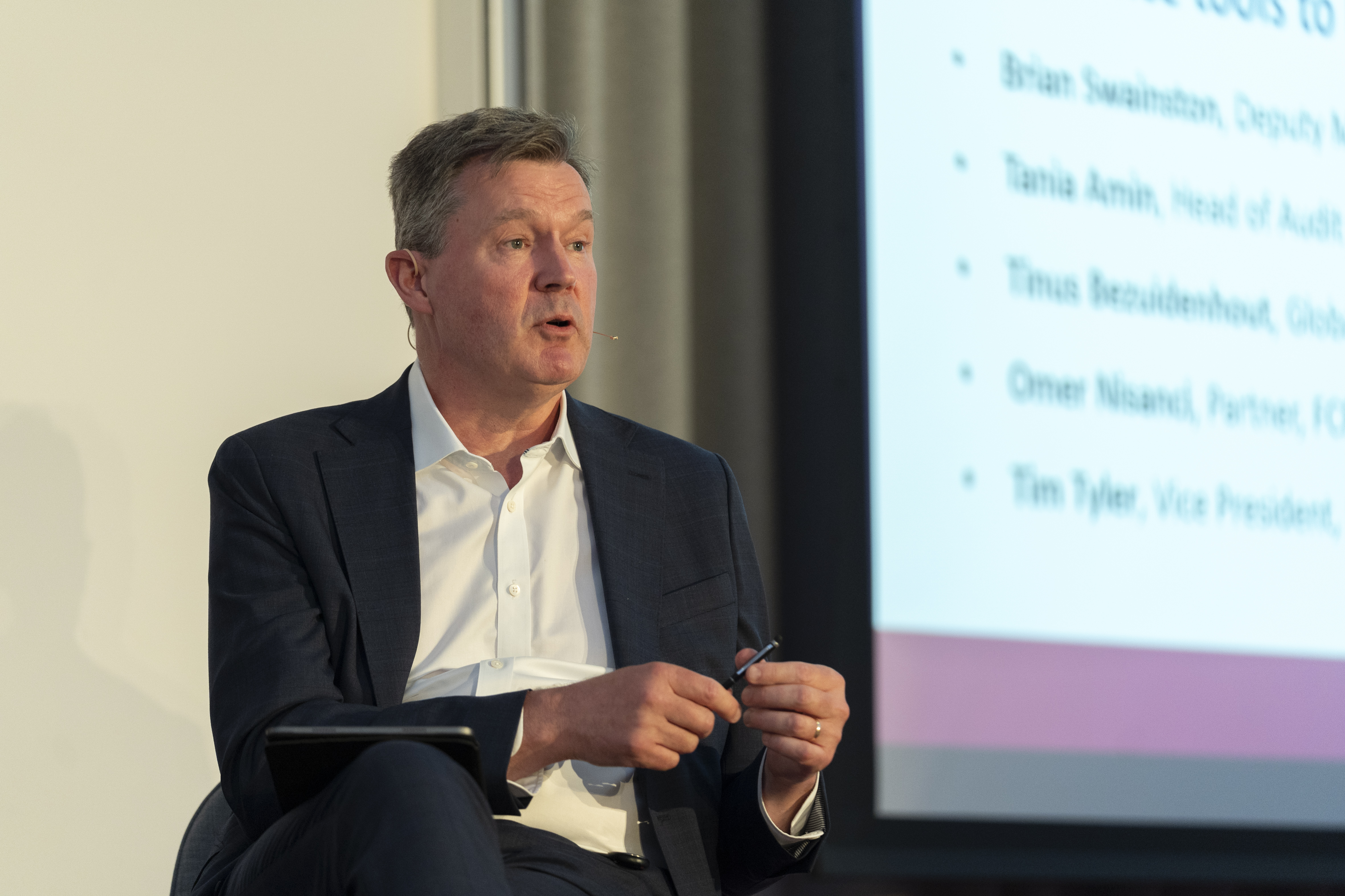 A man speaking at a conference. He is sat on a chair