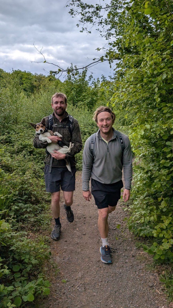Two men walking on a leaft path. One an is carrying a dog